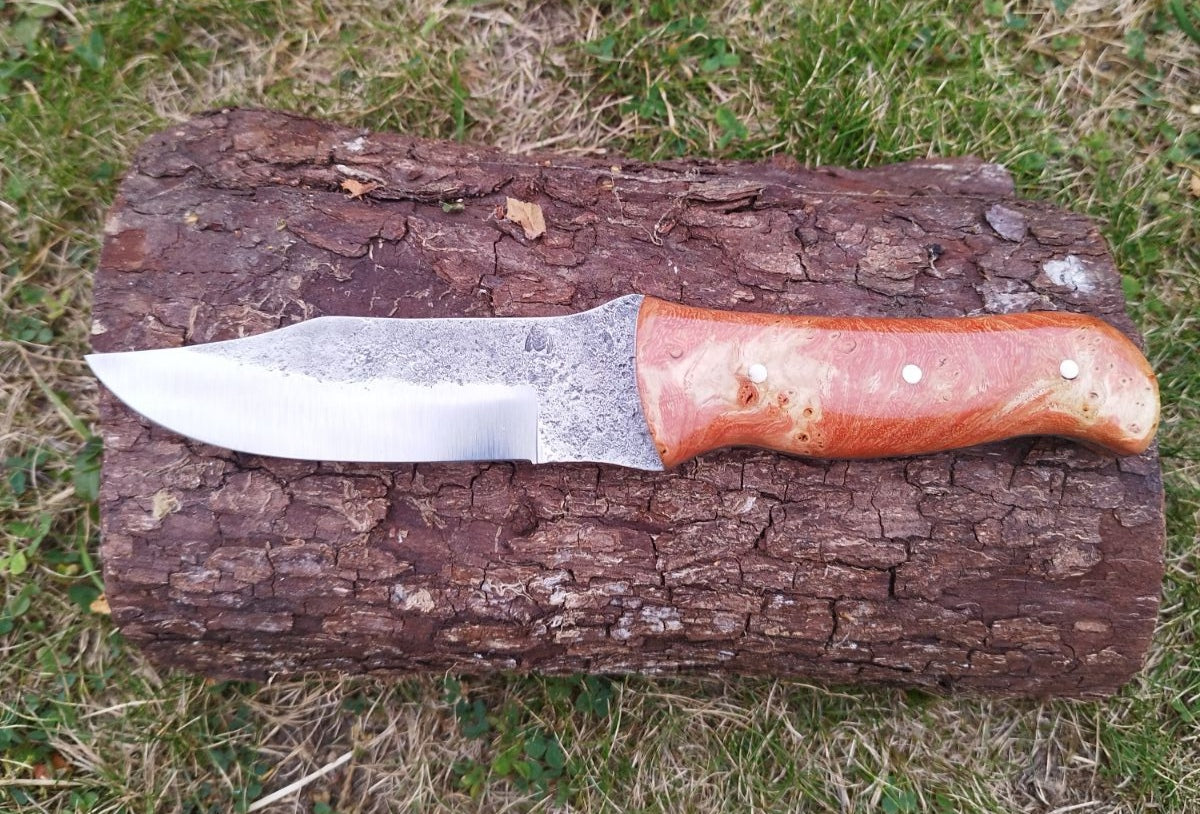 Créé en août 2024, ce couteau de survie avec un haut de lame courbé, brute de forge sur la lame et  un manche en loupe de peuplier stabilisé rouge